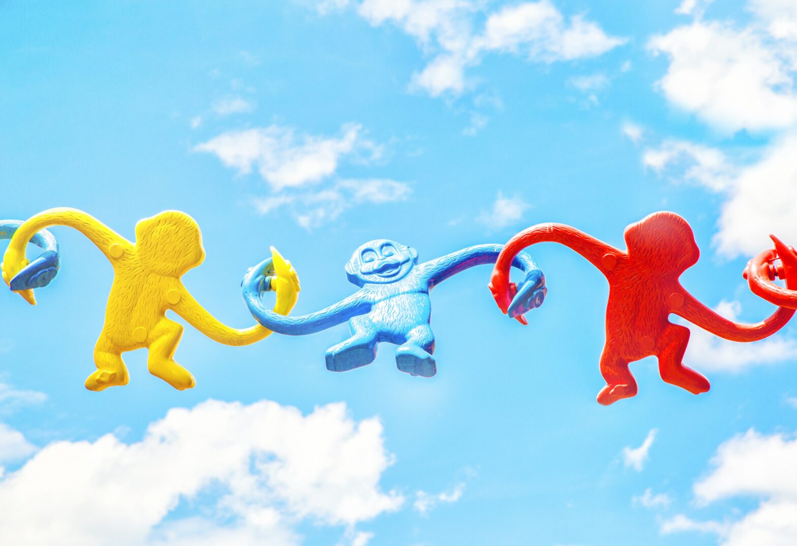 red yellow and blue monkeys interlinked by their arms in front of a blue sky with clouds